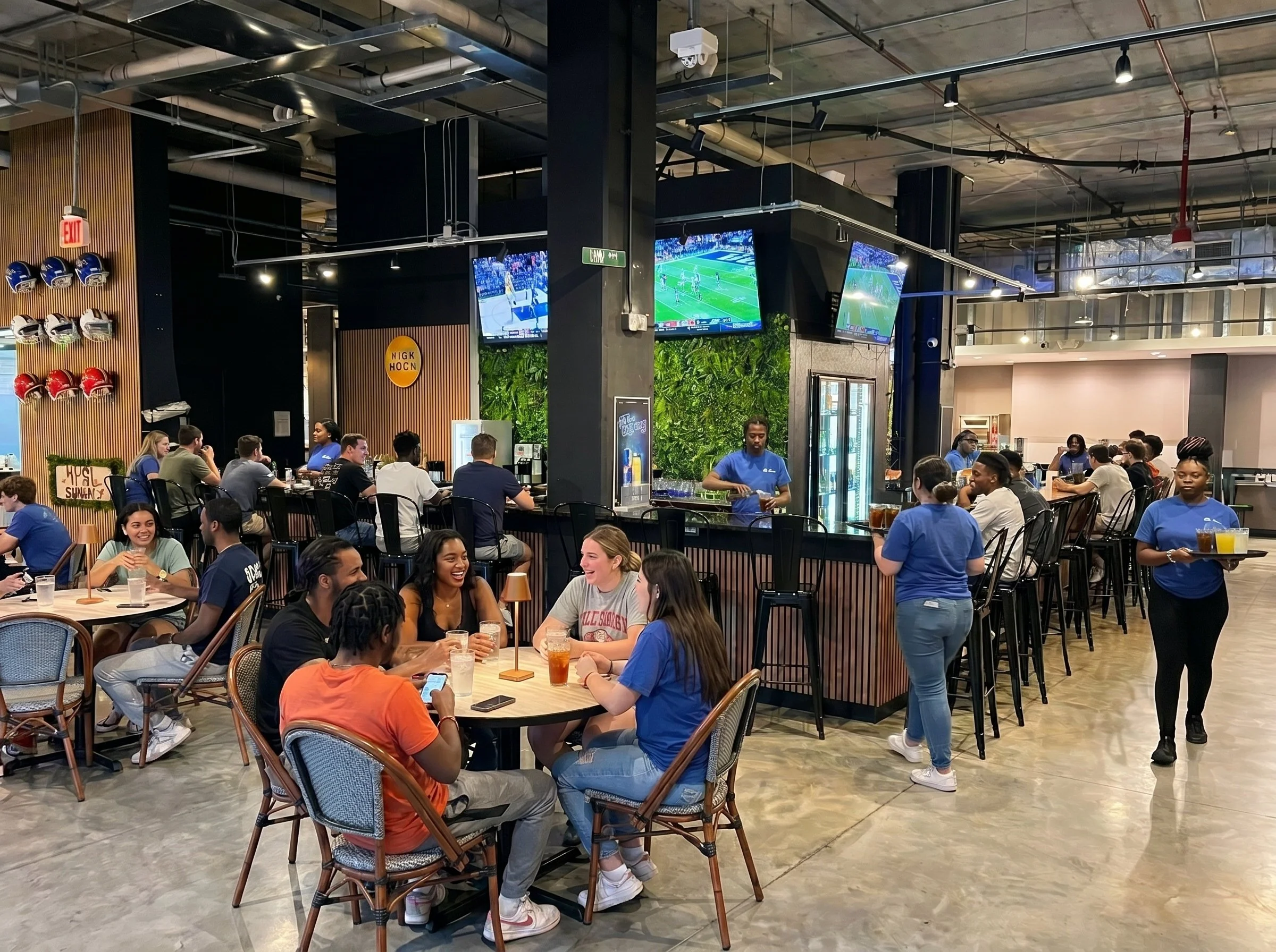 A lively indoor sports bar filled with people sitting at tables, watching multiple large TVs displaying a football game, with waitstaff serving drinks.
