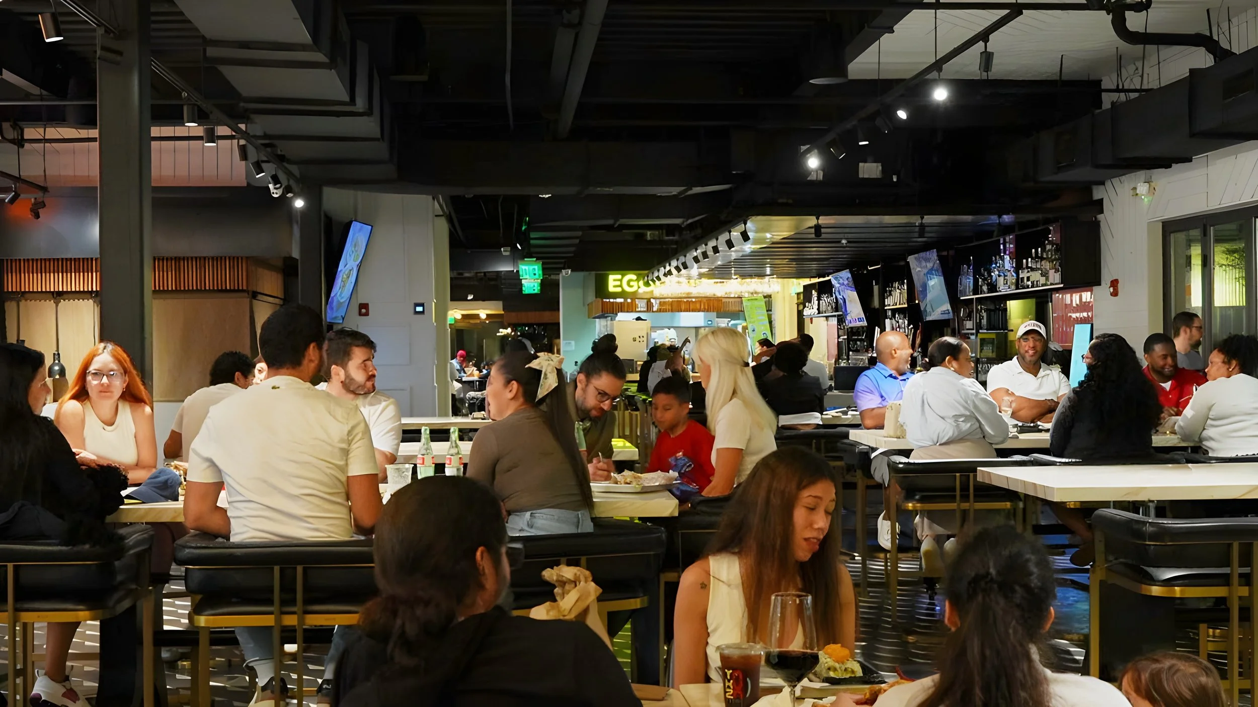Crowded indoor restaurant or food court with diverse group of people sitting at tables, eating, talking, enjoying meals in modern decor with digital screens and bar area in the background.